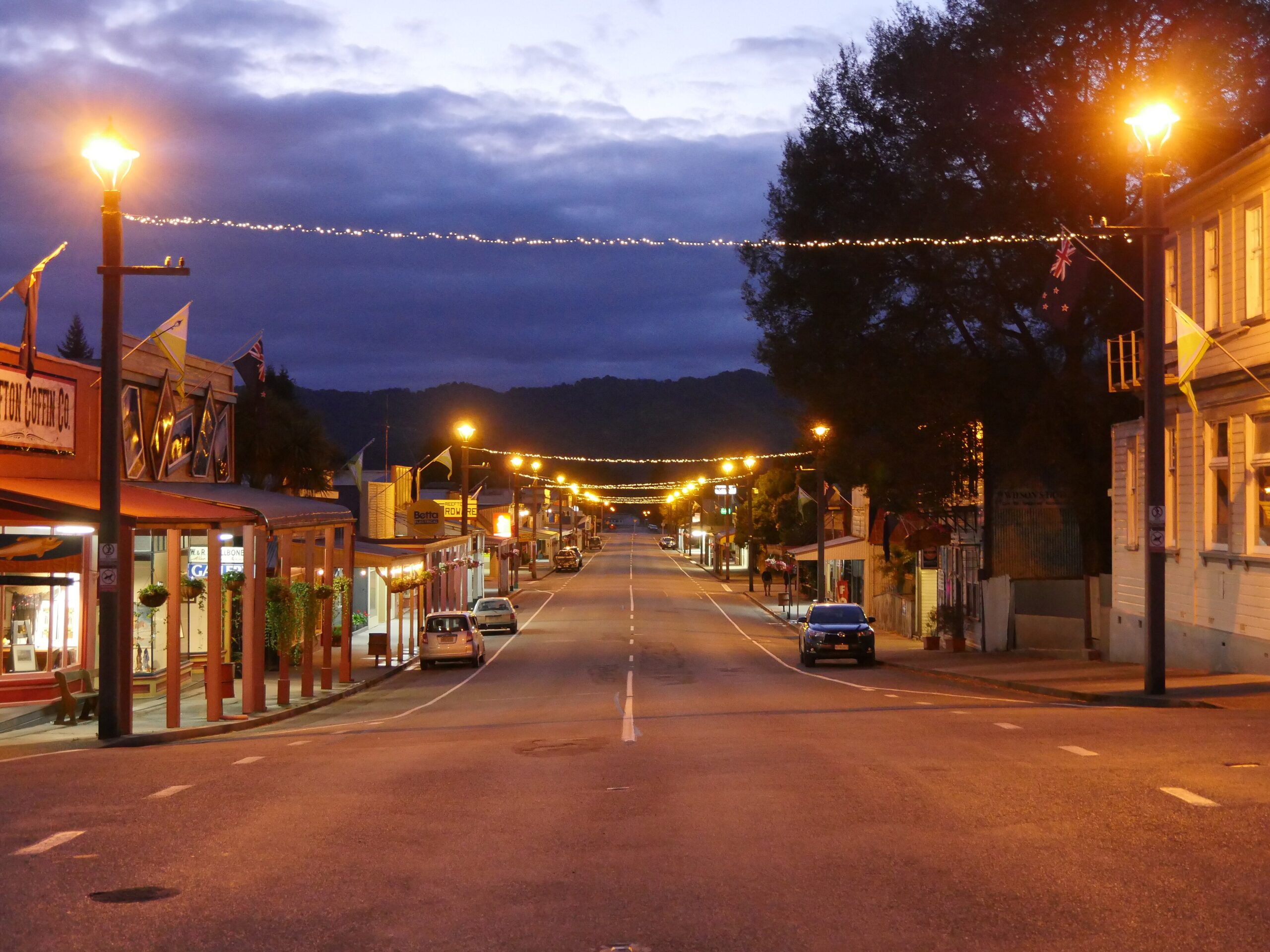 Broadway, the main street of Reefton, New Zealand, at dusk, with heritage-style street lamps and strings of lights over the road, timber shopfronts and bush-covered hills behind